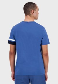 Blauwe katoenen T-shirt met korte mouwen, met een marineblauwe en witte gestreepte accent op de mouw en een klein rood, wit en blauw detail bij de hals.