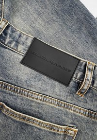 Blauwe denim jeans met een zwart geëmbosserd leren label met "BLOCKBINS." Zichtbare stiksels in contrasterend geel rond de tailleband.