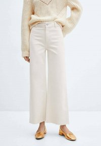 Pantalones de denims de pierna ancha color crema con dobladillo sin terminar, combinados con un suéter beige texturizado y zapatos tipo slip-on dorados.