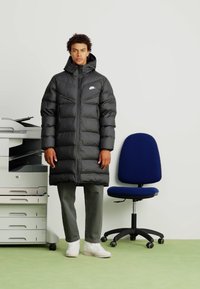 Crna puffer jakna s kapuljačom, s patentnim zatvaračem sprijeda i malim logotipom. Nosena na zelenim hlačama i bijelim tenisicama, protiv jednostavne pozadine.