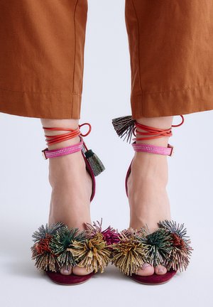 Pies con sandalias de punta abierta adornadas con borlas de cuentas multicolores y correas rojas en los tobillos, combinadas con pantalones marrones recortados.