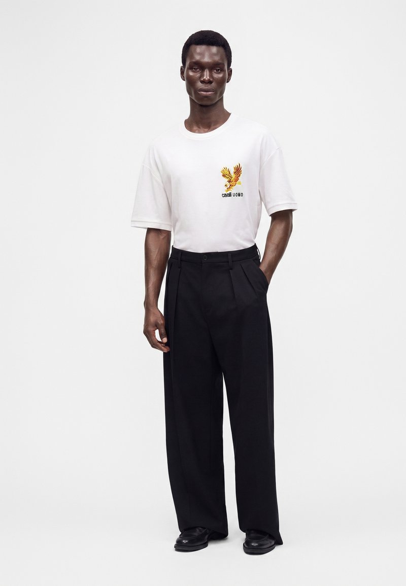 Hombre de pie con camiseta blanca de manga corta con diseño de águila amarillo-naranja y pantalones negros de pierna ancha sobre fondo blanco.
