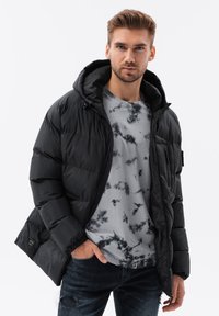 Schwarze Steppjacke mit Kapuze, ausgestattet mit einem Reißverschluss, elastischen Bündchen und einer strukturierten Oberfläche, getragen über einem grauen Batik-T-Shirt.