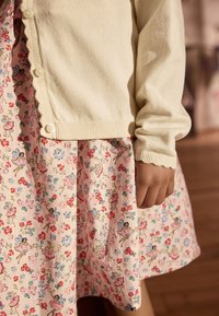 Light cream cardigan with scalloped edges, worn over a floral-patterned dress featuring pastel colors, ballet fairies, and small blossoms.