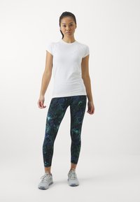 Maglietta sportiva bianca a maniche corte abbinata a leggins aderenti con un motivo a foglie tropicali verde scuro. Scarpe da ginnastica grigio chiaro.