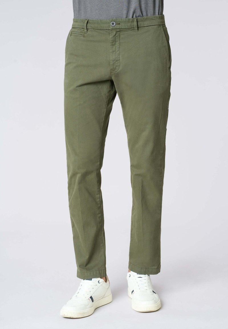 Pantaloni in cotone verde oliva con vestibilità dritta, fronte piatto e tasche laterali, abbinati a sneakers bianche. Texture del tessuto sottile visibile.