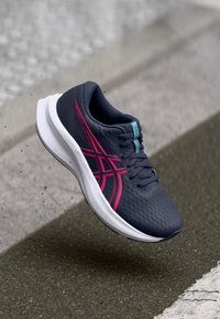 Navy blue running shoe with pink stripes and white sole, hovering above textured pavement near a white road marking.