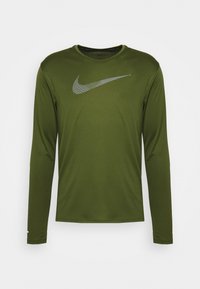 Olivenfarbiges langärmeliges Sportshirt mit einem großen grauen Nike-Swoosh-Logo über der Brust auf einem schlichten weißen Hintergrund.
