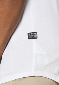 Valge puuvillane T-särk, millel on must ristkülikukujuline silt, millel on külje lähedal hemmi õmmeldud tekst "G-STAR ORIGINALS RAW DENIM".
