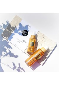 Två gyllene kroppssprayflaskor med trälock på en texturerad stonyta, bredvid ett vitt kort med en svart cirkulär klistermärke och text.