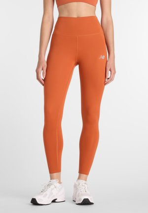 Γυναίκα που φοράει αθλητικό κολάν ψηλόμεσο σε καμένο πορτοκαλί χρώμα και λευκά αθλητικά παπούτσια New Balance, στέκεται μπροστά σε έναν απλό φωτεινό φόντο.