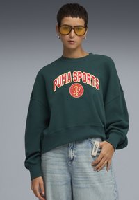 Zelená oversized mikina s logom "PUMA SPORTS" v červenej a žltej farbe. V kombinácii s svetlomodrými džínsami, ktoré majú dekoratívny korálkový doplnok.