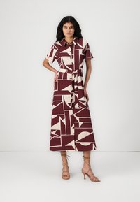 Robe chemise maxi en bordeaux avec des motifs géométriques beiges, manches courtes, fermeture à boutons sur le devant, et taille nouée. Associée à des talons beiges à lanières.