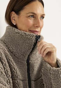Fleecejacke in hellem Beige mit hohem Kragen, Reißverschluss und strukturierten Oberflächen. Schwarze Reißverschlussdetails sichtbar.