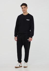 Felpa nera con collo tondo e polsini a maglia, abbinata a pantaloni jogger coordinati. Entrambi presentano un piccolo logo bianco sul petto e sulla coscia.