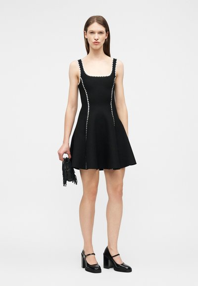 Femme en robe noire sans manches avec bordure blanche en zigzag tenant un petit sac noir à franges, portant des escarpins noirs de style Mary Jane, debout sur fond blanc.