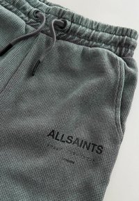 Záber zblízka na šedé tepláky s textúrou, elastickým pásom, šnúrkami, bočným vreckom a čiernym logom "ALLSAINTS" s menším textom pod ním.