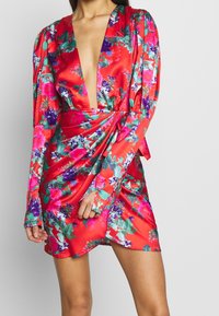 Robe en satin rouge à manches longues, décolleté en V profond, motif floral rose et violet, taille accentuée et design enveloppant.