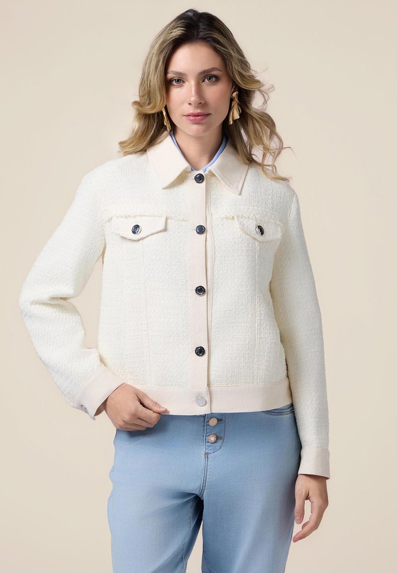 Giacca di tessuto crema con colletto in contrasto pallido, quattro tasche con bottone e bottoni scuri. Abbinata a pantaloni di denim azzurro chiaro.