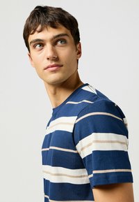 Camiseta a rayas con líneas horizontales en azul marino, blanco y beige, cuello redondo y mangas cortas, con una textura de algodón suave.