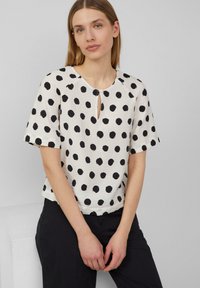 Haut blanc à pois noirs, encolure ronde, manches courtes et détail en forme de trou de serrure à l'avant. Le tissu a une finition texturée.