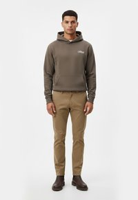 Sudadera marrón con bolsillo frontal y logo, combinada con pantalones beige y botas marrones oscuras. Diseño simple con un ajuste relajado.