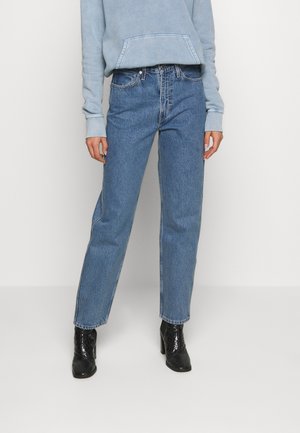 Personne portant un sweat-shirt bleu clair, un jean bleu taille haute à jambes droites, et des bottines à talons noires, debout contre un mur blanc.