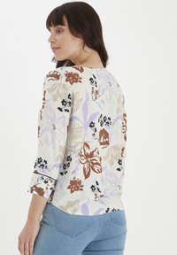Blusa floreale in crema con motivi in viola tenue, rosso e nero. Presenta maniche a tre quarti e un orlo arrotondato. Texture liscia.