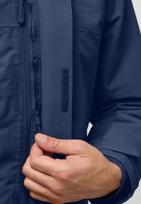 Giacca resistente all'acqua di colore blu navy con una superficie testurizzata, dotata di una cerniera frontale e dettagli in velcro; mano che tiene il tirante della cerniera.