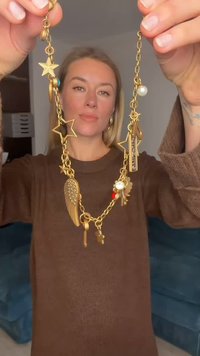 Liels zelta toņa kaklarota ar vairākiem piekariņiem, tostarp zvaigznes, spārni un pulkstenis, ar tekstūru un krāsu miksli.