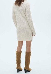 Personne portant une longue robe-pull en tricot torsadé crème et des bottes hautes en daim beige avec talons carrés, debout devant un fond uni.