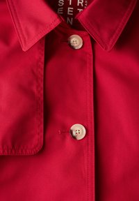 Červená látková košile s velkým límcem, bílým knoflíkem se čtyřmi otvory a diagonálními švy na přední straně.