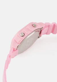 Montre en silicone rose avec un bracelet texturé, des boutons argentés et un dos métallique affichant des gravures. Elle a une forme rectangulaire.