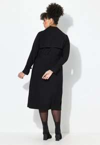 Schwarzes, langärmliges Kleid mit tailliertem Gürtel, Kragen und asymmetrischem Saum. Glattes Material und Schlitze an den Seiten. Getragen mit Stiefeletten.