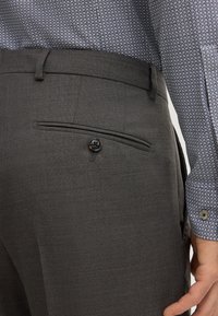 Gray textured trousers featuring a back pocket with a button closure. Accompanied by a patterned blue and white shirt sleeve.