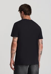 Czarna koszulka z krótkim rękawem wykonana z bawełny, z okrągłym dekoltem, luźnym krojem i prostym wzorem, z widocznymi tatuażami na ramionach modela.