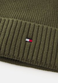 Căciulă de tricot rib, de culoare verde oliv, cu un mic logo brodat care prezintă dungi roșii, albe și albastre pe partea din față.