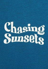 Modrá látka s výrazným bílým textem říkajícím "Hledání západů slunce" v vlněném písmu. Textura je hladká a konzistentní po celé ploše.