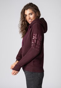 Bordeauxkleurige hoodie met een voorzak, capuchon met trekkoord en grote tekstafdruk op de mouw. Voorzien van ribgebreide manchetten en onderzoom voor een snug fit.