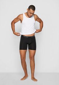 Fekete boxer alsónadrág elasztikus derékszíjjal, amelyen a "Calvin Klein" márkajelzés látható, fehér ujjatlan pólóval kombinálva egy modellen.
