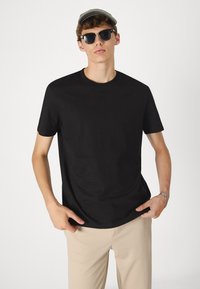 Sort T-shirt med korte ærmer, rund halsudskæring, båret sammen med beige bukser. Model iført grå kasket og sorte solbriller. Enkel design, blød tekstur.