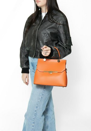 Borsa a mano in pelle arancione con una forma strutturata, manico superiore, accessori in oro e chiusura a pattina. Il modello indossa una giacca bomber nera e jeans.