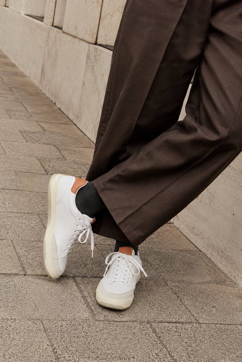 Person trägt weiße Sneakers mit braunen Akzenten, dunkle Socken und braune Hosen und steht auf einem gefliesten Gehweg in der Nähe einer Steinmauer.