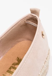 Gros plan sur une espadrille beige montrant l'intérieur en daim, la semelle intérieure en cuir avec logo, la semelle en jute tissée, et une languette de tirage au talon.