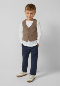 Brązowa kamizelka w kratę z czterema guzikami, noszona na białej koszuli z długim rękawem, zestawiona z granatowymi spodniami i beżowymi sneakersami. Prosty design.