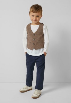 Brauner Tweed-Weste mit vier Knöpfen, getragen über einem weißen Langarmhemd, kombiniert mit blauen Hosen und beigen Sneakers. Einfaches Design.