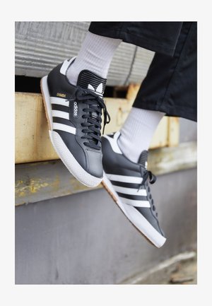 Fekete Adidas Samba sportcipők fehér csíkokkal, textúrált gumitalppal és fekete bordázott bokakollárral. Fehér bordázott zoknival viselve.
