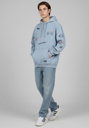 Felpa azzurra chiaro con vari ricami, tra cui ali e testo; abbinata a jeans azzurri chiari e sneakers bianche con strisce nere.