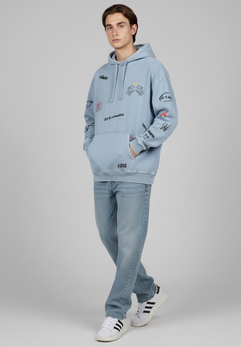 Sweat à capuche bleu clair avec divers motifs brodés, y compris des ailes et du texte ; associé à un jean bleu clair et des baskets blanches avec des rayures noires.