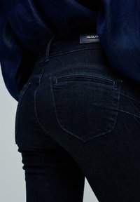 Jeans de mezclilla azul oscuro con un ajuste ceñido, que presentan detalles de costura sutiles y dos bolsillos traseros. La cinturilla tiene una etiqueta de marca.
